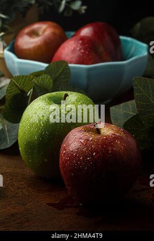 Wet green and red apples placed on wooden table against blurred background of bowl with red apples near leaves Stock Photo