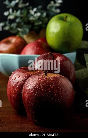 Wet red apples placed on wooden table against blurred background of bowl with red and green apples near pot Stock Photo