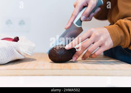 Crop anonymous female halving ripe black avocado with knife on chopping board while cooking in kitchen at home Stock Photo