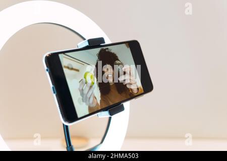 From above of smiling African American female blogger recording video about cooking healthy food on cellphone using ring lamp against white wall Stock Photo