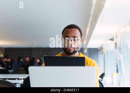 Thoughtful black male sitting at table with laptop and listening for music in earphones from tablet in coworking space Stock Photo