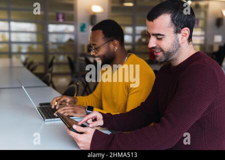 Smiling multiracial bearded partners sitting together at table with laptop and tablet in coworking center Stock Photo