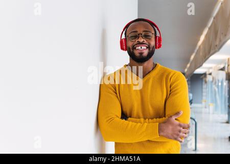 Relaxed smiling bearded African American male in sweater and eyeglasses looking at camera enjoying music in wireless headphones while leaning on wall Stock Photo