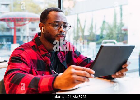 Concentrated African American bearded man in checkered shirt and eyeglasses browsing tablet with earphones while resting in cafe and looking at screen Stock Photo