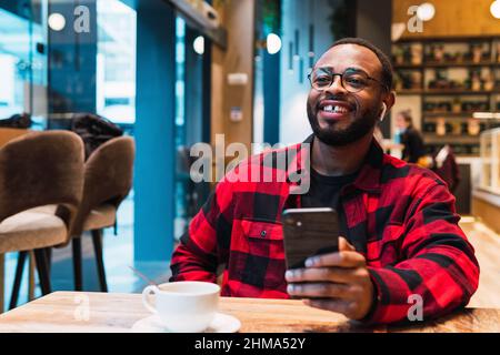 Smiling African American bearded male in red checkered shirt messaging on mobile phone wit earphones while drinking coffee in cafe and looking away Stock Photo