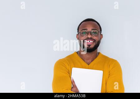 Positive bearded African American male in yellow sweater and eyeglasses holding notebook while looking at camera and standing against white background Stock Photo