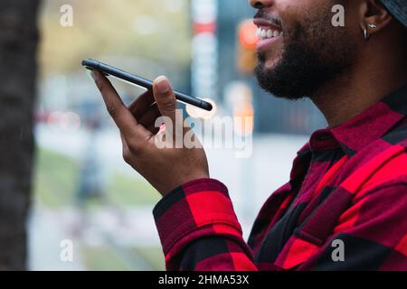 Happy cropped unrecognizable bearded African American male in checkered shirt and hat recording voice message on mobile phone in city street Stock Photo