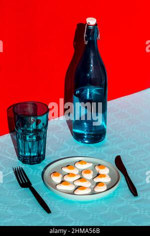 From above of composition of bottle of water with glass opposite plate with halved boiled eggs and fork and knife placed on blue surface against red b Stock Photo