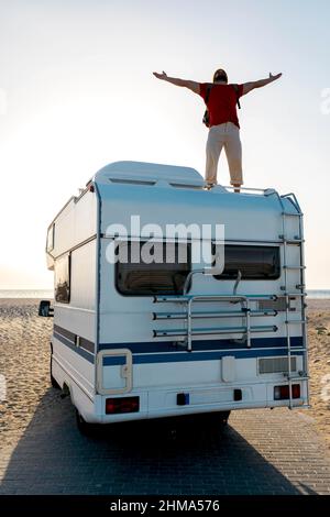 Carefree male explorer standing on roof of camper parked on seashore and enjoying freedom with outstretched arms against sundown sky Stock Photo