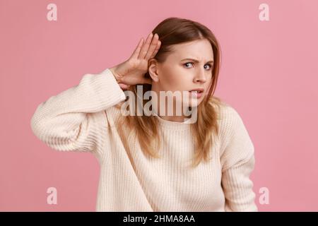 Portrait of blond woman holding hand near ear and listening carefully, having hearing problems, deafness in communication, wearing white sweater. Indoor studio shot isolated on pink background. Stock Photo