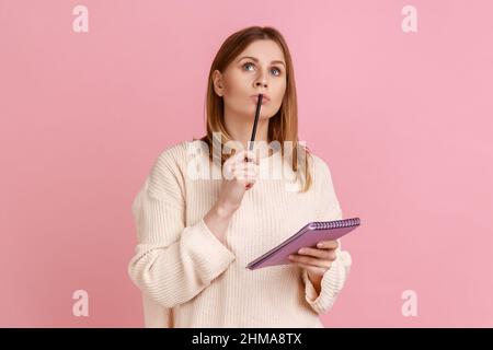Portrait of pensive blond woman holding pen and paper notebook, being deep in thoughts, writing a composition, wearing white sweater. Indoor studio shot isolated on pink background. Stock Photo