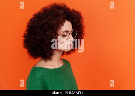 Profile portrait of sad upset woman with Afro hairstyle wearing green casual style sweater standing with pout lips, feels sadness, cry. Indoor studio shot isolated on orange background. Stock Photo
