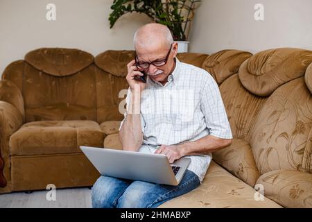 Smiling mature 60s caucasian man looking at laptop screen and talking on the phone. A serious gray-haired old man makes an order in an online store. U Stock Photo