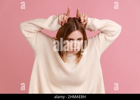 Portrait of aggressive blond woman making bull horns gesture over her head and frowning face, expressing anger threat, wearing white sweater. Indoor studio shot isolated on pink background. Stock Photo