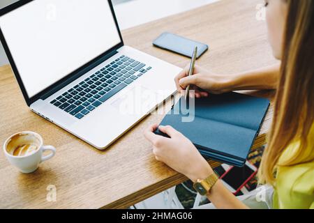 Unrecognizable lady working on laptop while making notes in notepad Stock Photo