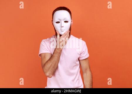 Portrait of unknown bearded man covering her face with white mask, hiding her real personality, anonymity, wearing pink T-shirt. Indoor studio shot isolated on orange background. Stock Photo