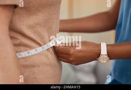 Just the weight its supposed to be. Shot of a doctor using a measuring tape on a senior womans stomach during a consultation at home. Stock Photo
