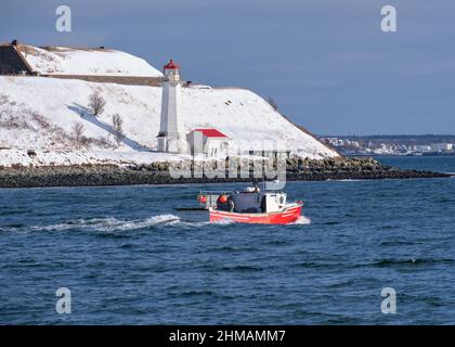 Red fishing boat passing by the Georges Island Lighthouse covered in snow on sunny winter day. Halifax, Canada Stock Photo