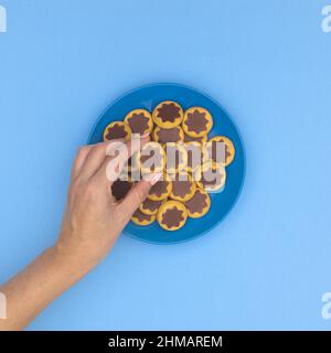 Woman hand taking one delicious chocolate and biscuit cookie from blue plate on cyan background. Food flat lay. Stock Photo