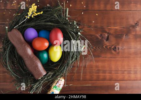 Easter eggs and chocolate bunny in a nest of branches on dark wooden background. Copy space. Top view. Stock Photo