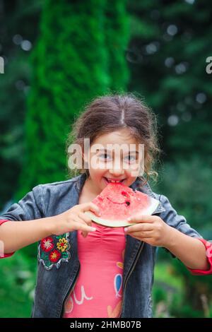 Little happy girl eating a juicy watermelon in the garden. Children eat fruit outdoors. Healthy food for children. Stock Photo
