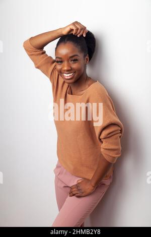 A beautiful black woman standing against a white wall with one hand held near her head. Stock Photo