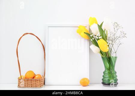 Composition with photo frame, Easter eggs in basket and flowers on table near light wall Stock Photo