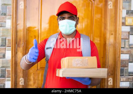 Focus on face, delivery boy with medical face mask and cardboard boxes in hands showing thumbs up gesture bylooking at camera - concept of online Stock Photo