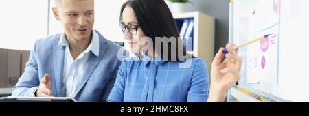 Young man and woman looking at screen of digital tablet and studying information on blackboard Stock Photo