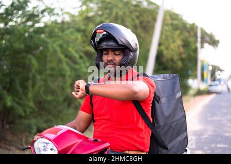 Worried Indian deliveryman on scooter due delayed delivey service - concept of stressful work lifestyle, employment and courier service. Stock Photo