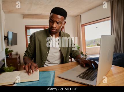 Young adult black male concentrating taking notes from laptop. Working from home, sitting at modern styled kitchen counter. Stock Photo