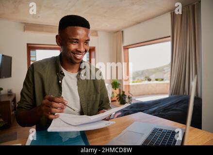 Happy young adult black male studying with paper notes, book and laptop in modern apartment.  Stock Photo