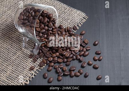 Coffee beans scattered from a glass cup on a black wooden surface with sacking Stock Photo