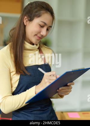Portrait of a beautiful asian female waitress taking a food and drink orders on clipboard paper. Friendly server wears apron and takes orders in resta Stock Photo