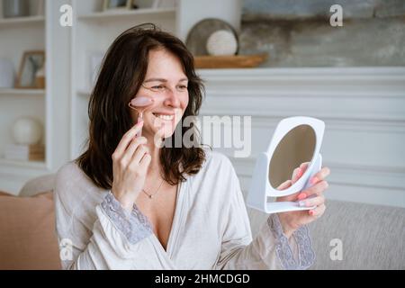 Woman massages her face with pink stone roller, facial massage at home, spa facial treatment. Redhead girl sits on the couch in a bathrobe and uses a facelift kit looks through the mirror Stock Photo