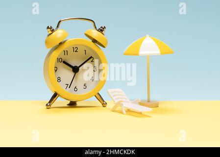 Time to travel. Sun lounger, yellow umbrella and alarm clock on a yellow, blue background. Stock Photo