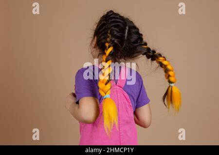 Back view of little excited girl with kanekalon pigtails of yellow color, jumping and playing wearing pink jumpsuit and purple t-shirt on beige Stock Photo