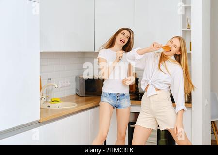 Two joyful women singing in the kitchen, dancing and pretend to be a rock star Stock Photo