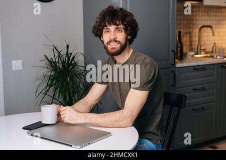 Portrait of cheerful curly young freelancer programmer male holding in hand cup with morning coffee sitting at table with closed laptop computer. Stock Photo