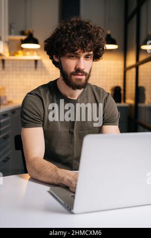 Vertical shot of focused bearded young freelancer male using typing on laptop keyboard, writing email or message sitting at table in kitchen. Stock Photo