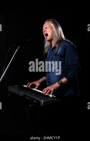 elderly man with long gray hair in blue denim shirt plays the keys and sings with microphone on stage on black background Stock Photo