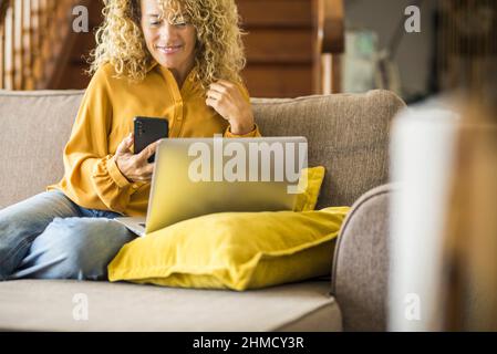 Excited and happy adult woman at home use mobile smartphone and laptop computer connection sitting on the sofa. Indoor leisure activity with wireless Stock Photo