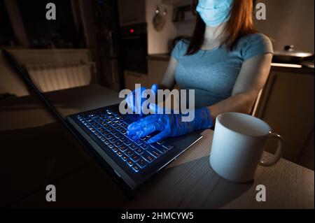 A faceless girl in a medical mask and glasses studies late in the kitchen during quarantine. Unrecognizable woman working remotely from home on a Stock Photo