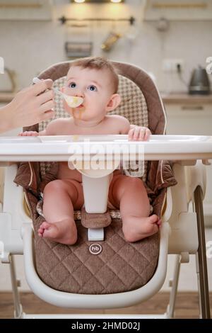 Mother feeding toddler baby from a spoon on a high chair for children, home kitchen background. Happy Baby (HB) - Moscow, Russia, January 26, 2022