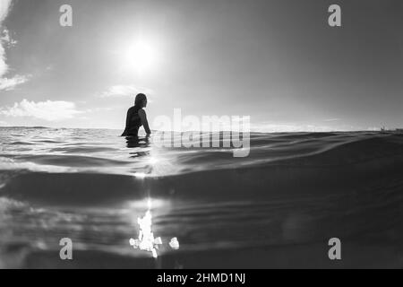 Surfing girl surfer silhouetted sitting on surfboard waiting in sunlight reflecting ocean water unrecognizable female rear behind  black and white pho Stock Photo