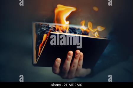 The man holds in his hand a black open book, the pages of which burn with a bright flame and smoke. The Antichrist burns a religious book. Censorship. Stock Photo