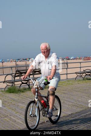 A senior citizen, apparently with a prosthetic lower leg. Riding a bicycle on the boardwalk in Brighton Beach, Brooklyn, New York City. Stock Photo