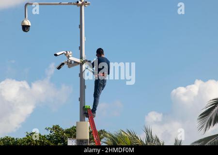 A male worker on a metal ladder installs security cameras near a public beach in Mexico Stock Photo