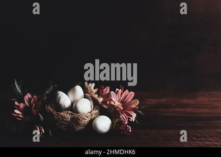 Easter holiday card with copy space to place text, mockup, wooden table background. White dotted Easter eggs in nest, pink flowers, bird feathers on wooden table. Minimal flat lay. Photo in low key Stock Photo