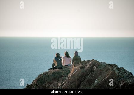 silhouette of three female hikers sitting on a rock and looks at sea with natural bokeh in soft warm sunset light. Selective Focus. Sea sunset Stock Photo